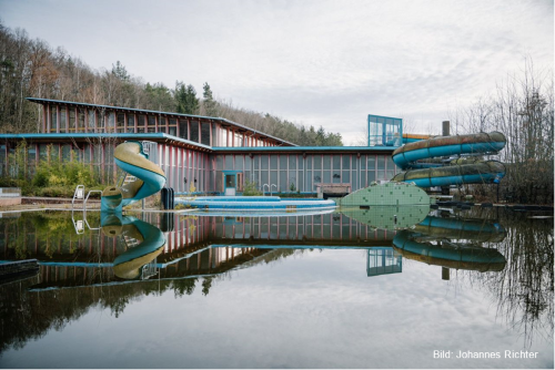 Verlassenes Schwimmbad mit Wasserrutschen und trübem Außenbecken im Wald.