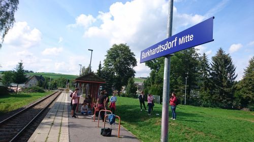 Personengruppe am Bahnsteig der Haltestelle Burkhardtsdorf Mitte bei sonnigem Wetter im Grünen.